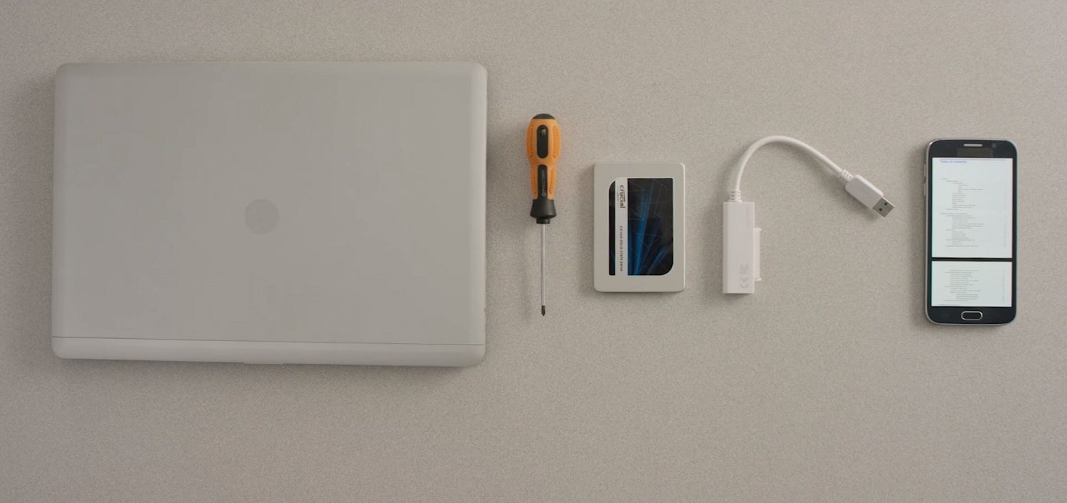 A laptop, Crucial SSD, screwdriver and computer’s owner’s manual on a mobile are laid out on a desk surface